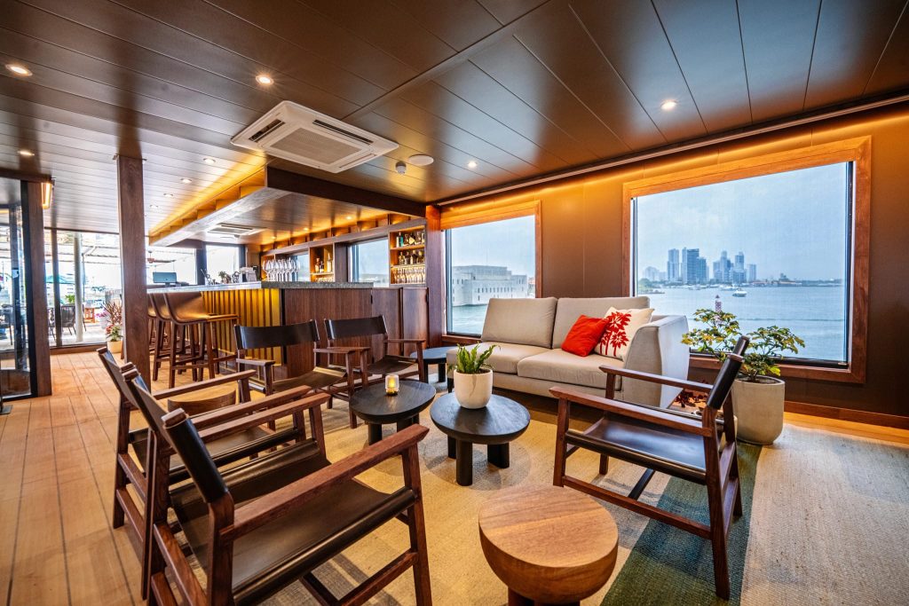 Lounge with bar, sofas and armchairs beside large windows overlooking the waterfront and city skyline.