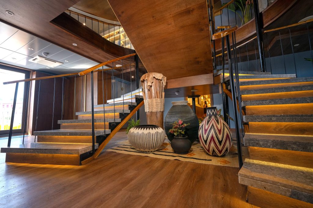 Ship lobby with a wooden staircase, glowing lit steps and large decorative woven and painted vases.