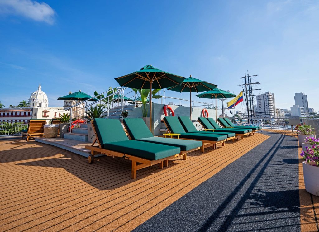 Sun deck with green-cushioned loungers under umbrellas, a colonial-era cathedral dome and a tall ship beyond.