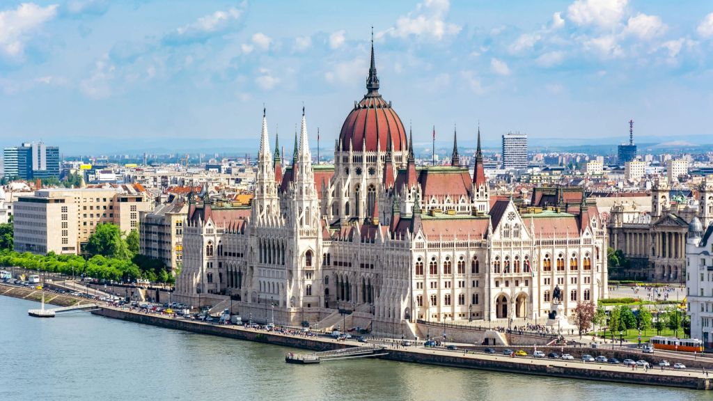 Budapest Parliament Building along the Danube with a red domed roof and ornate white facade under a blue sky.