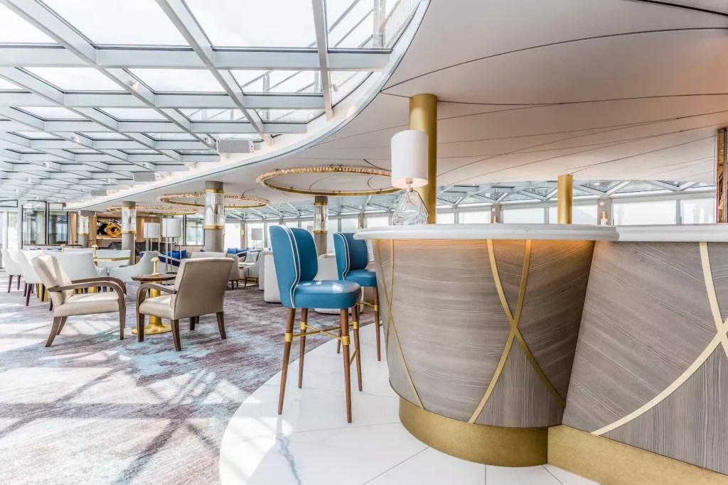 Ship lounge bar with a curved wood-and-gold counter, blue bar stools and a window-lined ceiling.
