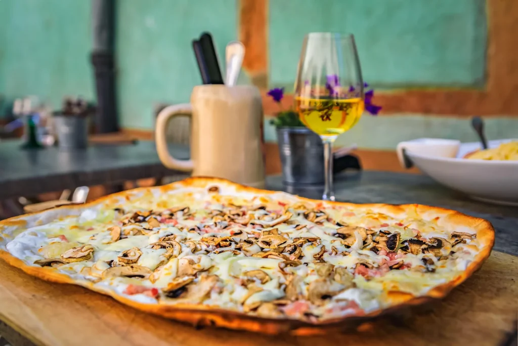 Thin-crust flatbread with mushrooms and cheese beside a glass of white wine on a wooden board.
