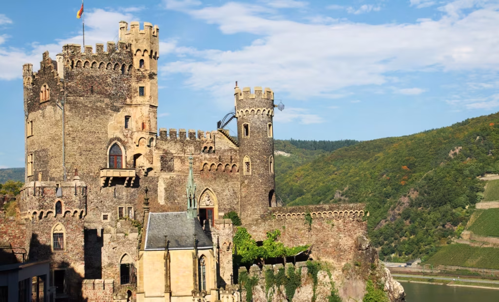 Medieval hilltop castle with towers and battlements above a river valley, lit by sunlight.