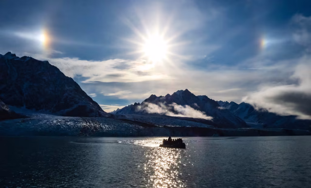 Small boat silhouetted on sunlit water beneath glacier-capped mountains with sun halos in the sky.