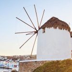 White windmill with a thatched roof on a stone base atop a grassy hillside, overlooking a white-washed coastal town.
