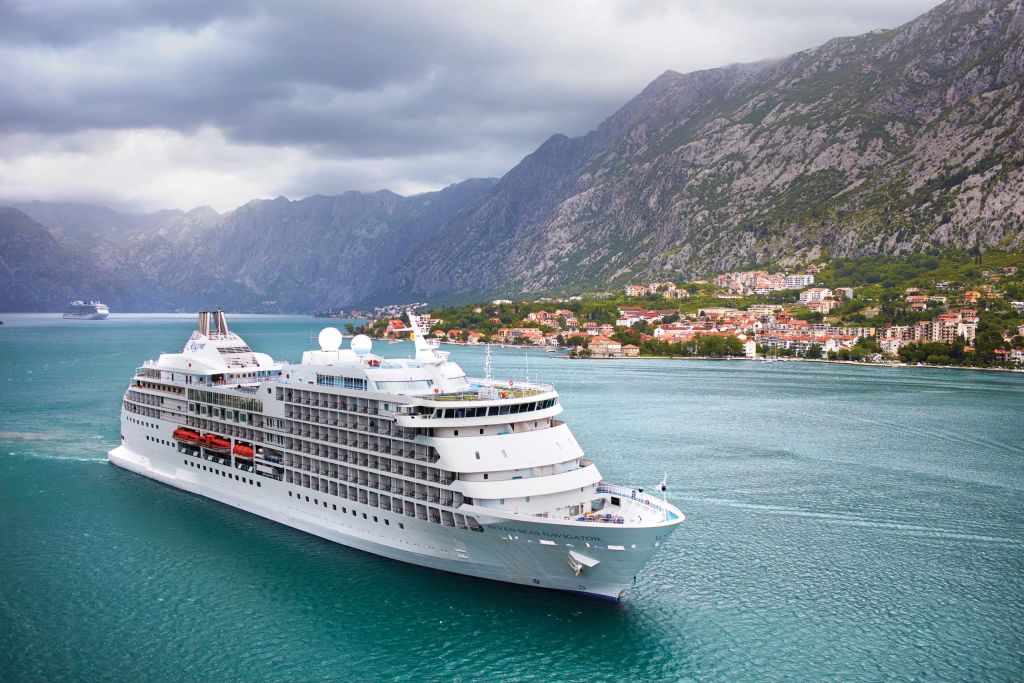 Cruise ship in a bay beneath steep mountains with a red-roofed hillside town.