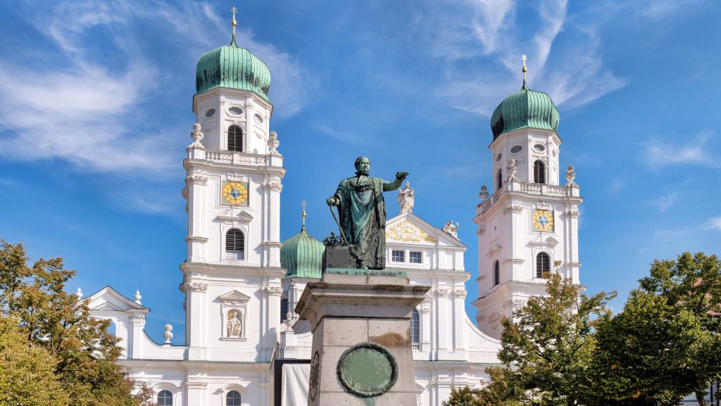 White Baroque church with two green onion domes and a bronze statue in front against a bright blue sky.