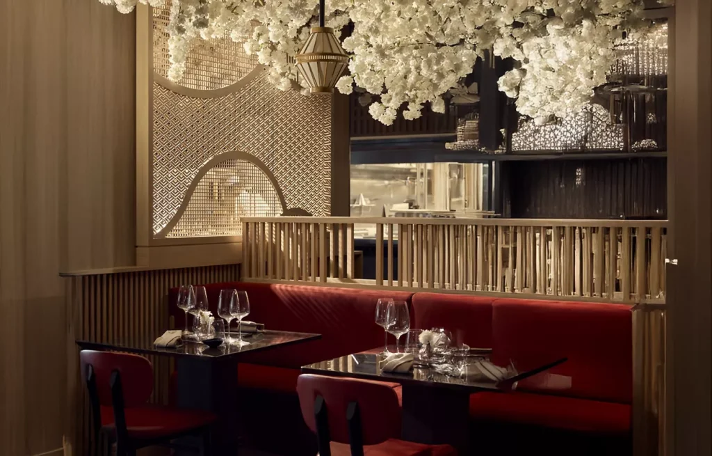 Dimly lit restaurant booth with red banquettes, white blossoms overhead and set tables.