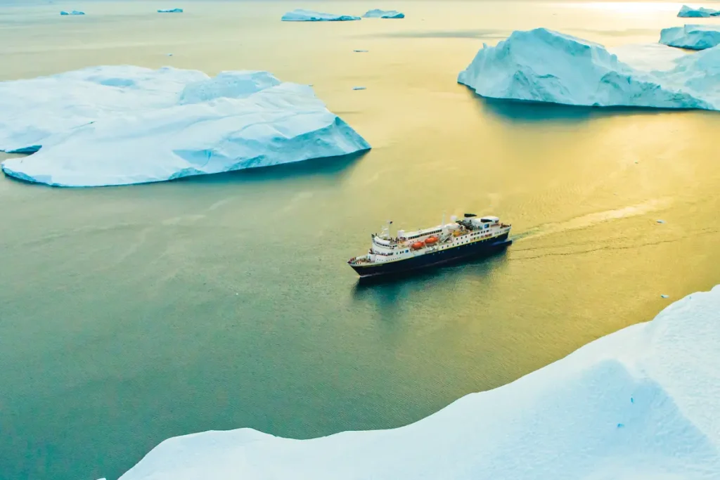 Small expedition ship sailing among large icebergs in golden evening light.