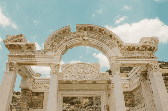 Ancient stone arch with carved frieze and supporting columns against a blue sky and clouds.