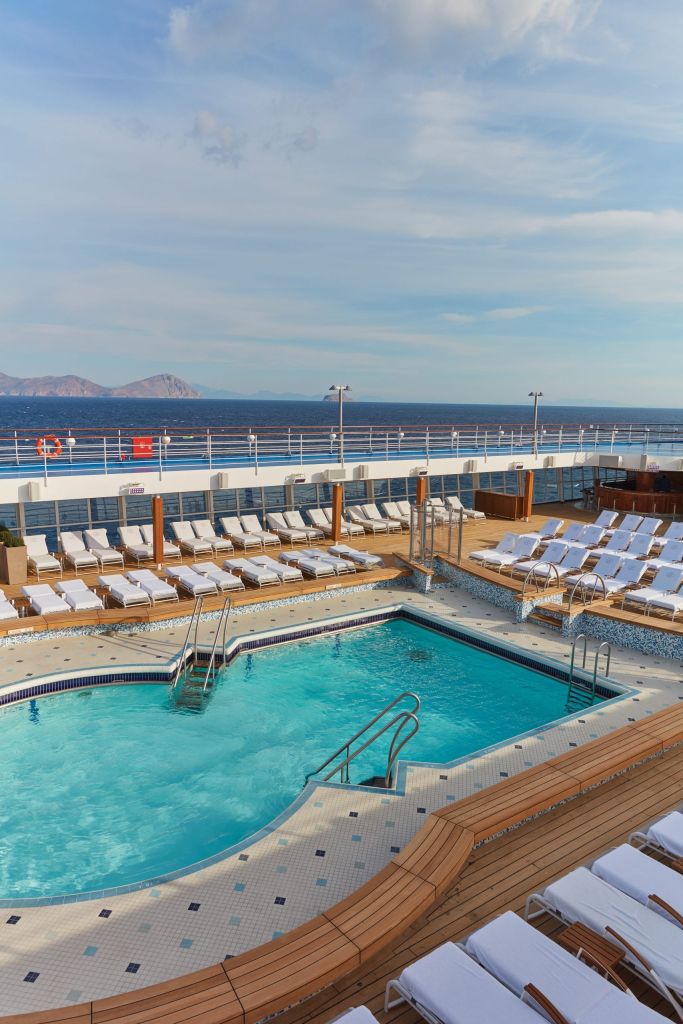 Ship pool deck with a blue swimming pool and rows of white loungers on wooden decking.