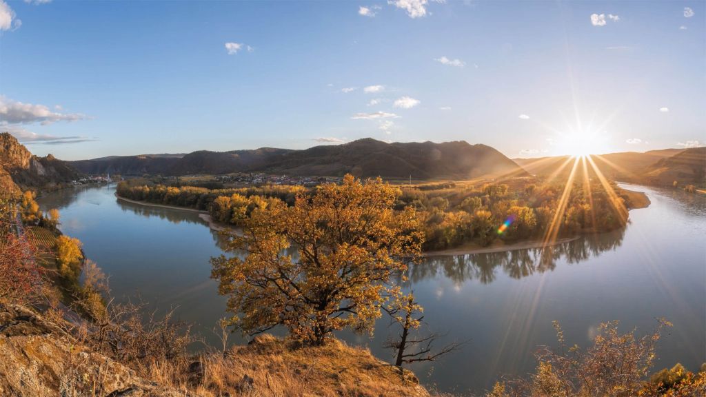 Wide river valley at sunset with golden autumn foliage and sunburst over hills in the distance