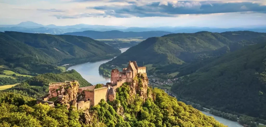 Ruined medieval castle on a rocky outcrop above a winding river valley with forested hills.