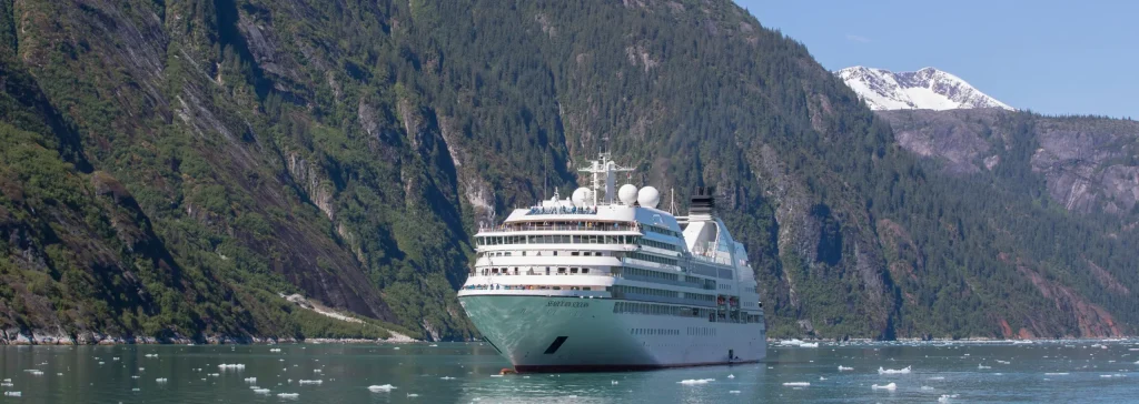 White cruise ship anchored in a fjord between steep green mountains, with floating ice.