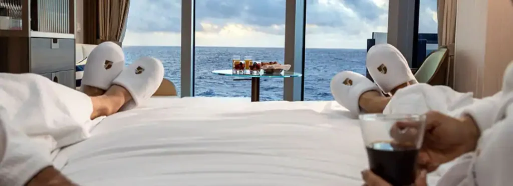 Two guests in white robes and slippers relax on a bed overlooking the ocean, one holding a drink.