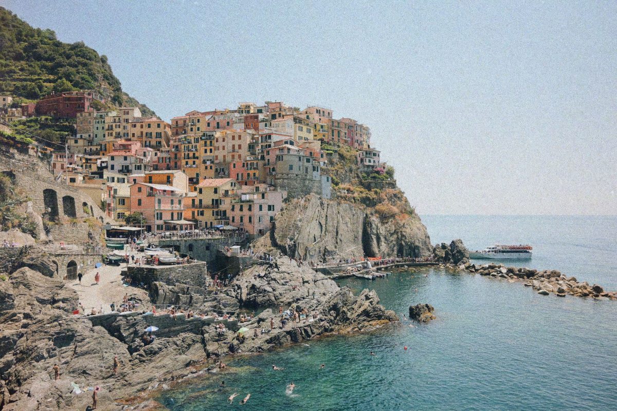 Colorful hillside village built into rocky cliffs above a calm harbor, with pastel houses and people along the rocky shore.