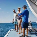 Couple in snorkel gear smiles on a boat deck while a crew member helps them prepare.