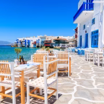 Outdoor cafe terrace with wooden chairs and tables overlooking whitewashed buildings and the sea.
