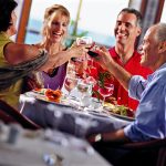 Four people in red and blue toast with wine glasses at a dining table by the window.