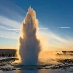 Geyser erupting into the air at sunset with spectators gathered behind.