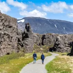 Hikers on a gravel path between dark basalt cliffs in a rift valley.