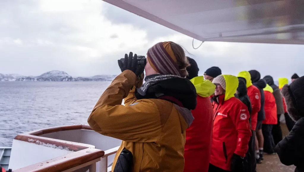 Guests in warm jackets and gloves watch through binoculars from a ship deck in cold weather.