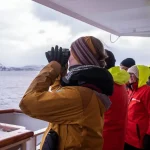 Guests in warm jackets and gloves watch through binoculars from a ship deck in cold weather.