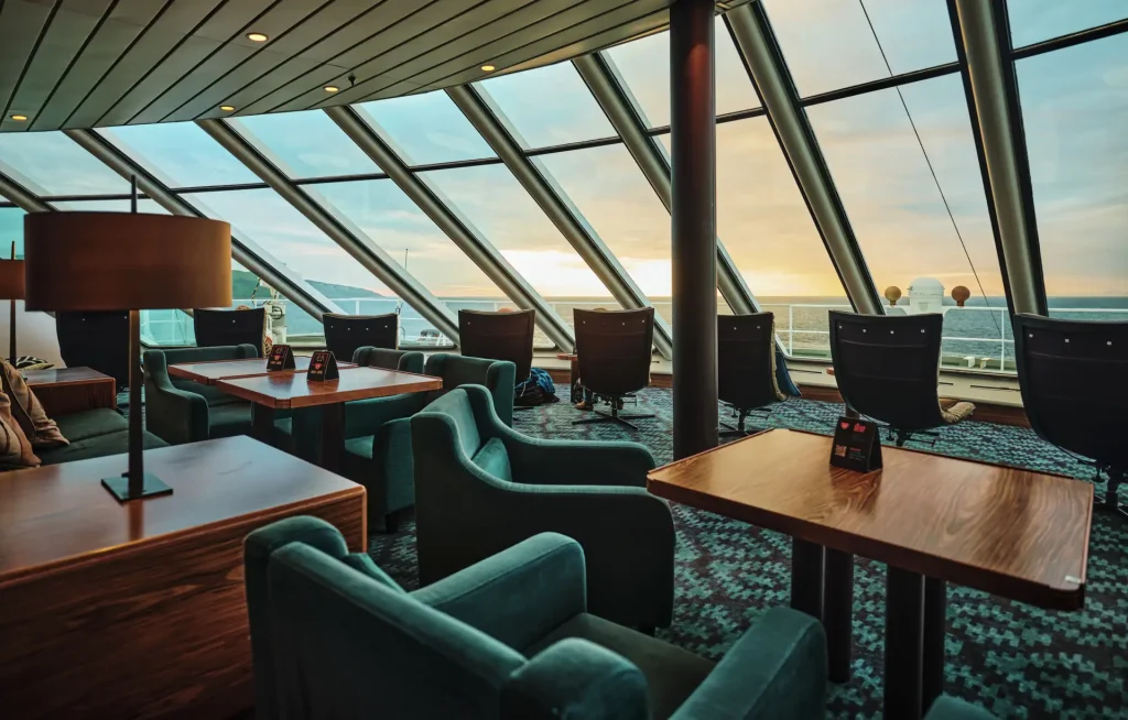 Ship lounge with angled glass windows, teal armchairs and wooden tables at sunset.