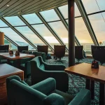 Ship lounge with angled glass windows, teal armchairs and wooden tables at sunset.