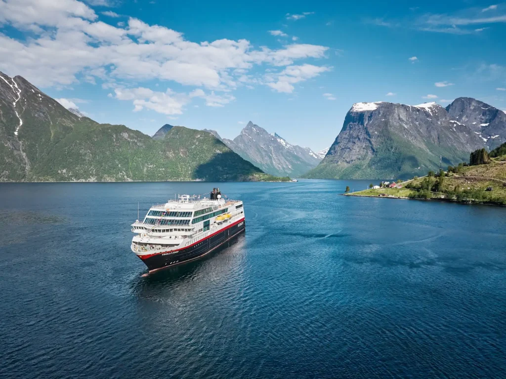 Red-and-white expedition ship sailing a deep blue fjord between green mountains.