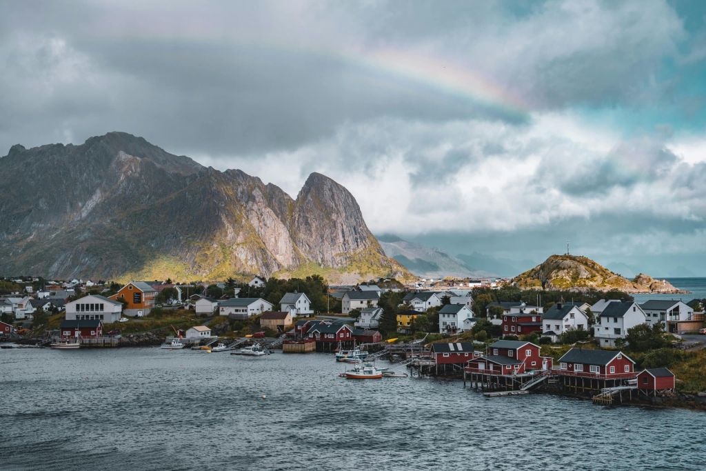 Red and white fishing village with houses on stilts beside a bay under mountains and a rainbow.