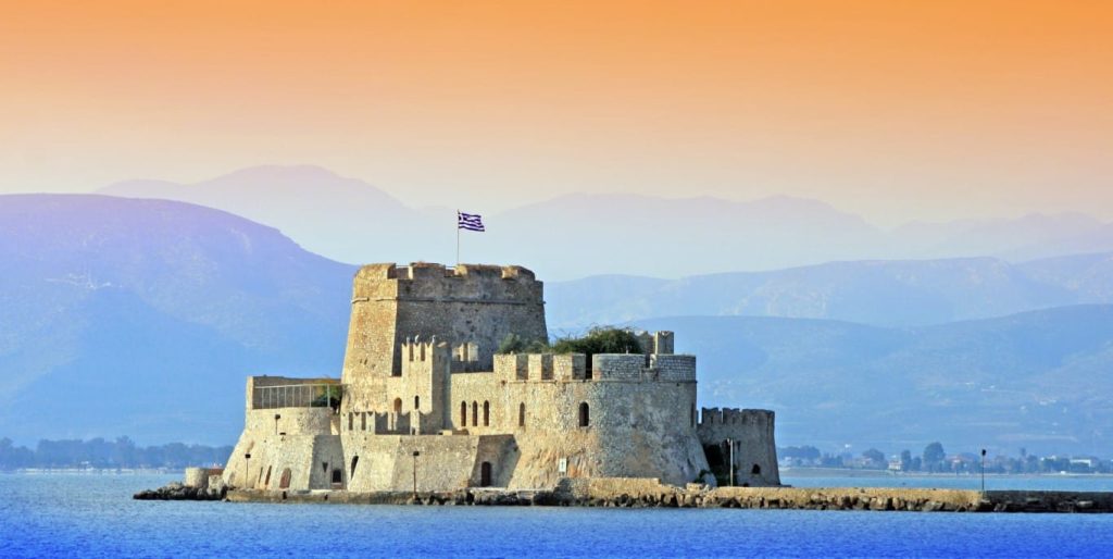 Sandstone sea fortress on a small island with a Greek flag, backed by hazy mountains at sunset.