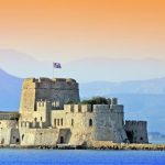 Sandstone sea fortress on a small island with a Greek flag, backed by hazy mountains at sunset.