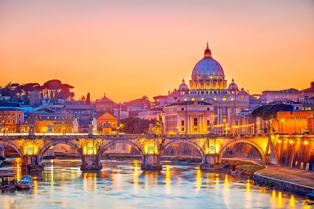 St Peter's Basilica dome glowing at sunset above a lit arched bridge over the Tiber in Rome.