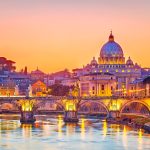 St Peter's Basilica dome glowing at sunset above a lit arched bridge over the Tiber in Rome.