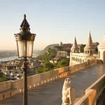 Stone terrace with ornate turrets and a classic street lamp overlooking a panoramic city view at sunset.