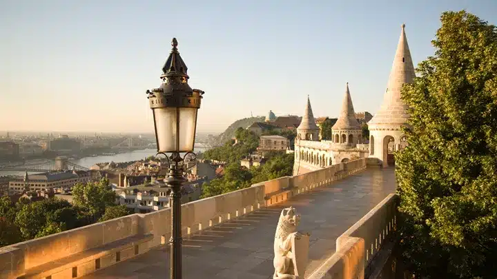 Stone terrace with ornate turrets and a classic street lamp overlooking a panoramic city view at sunset.