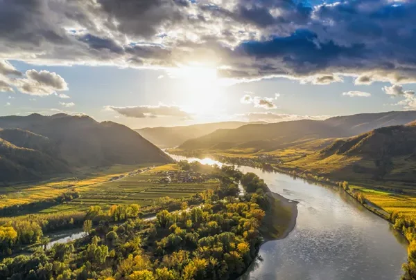 Sunlit river winding through a green valley with fields and rolling hills under a bright sky.