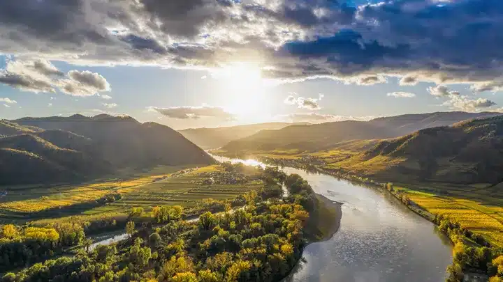 Sunlit river winding through a green valley with fields and rolling hills under a bright sky.