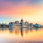 Budapest Parliament Building along the Danube at sunset, reflected in calm water.