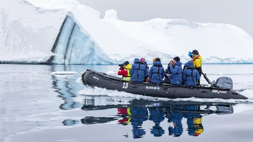 Passengers in blue jackets ride a black zodiac boat past a large iceberg.