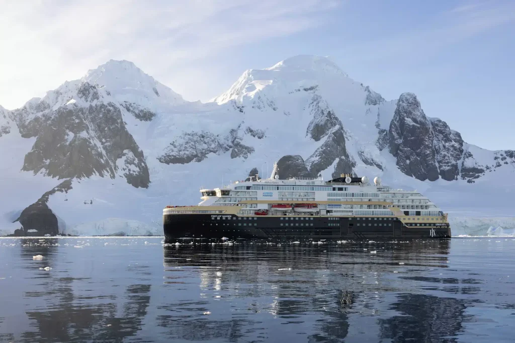 Expedition ship sails past snowy peaks and glaciers in calm polar water.