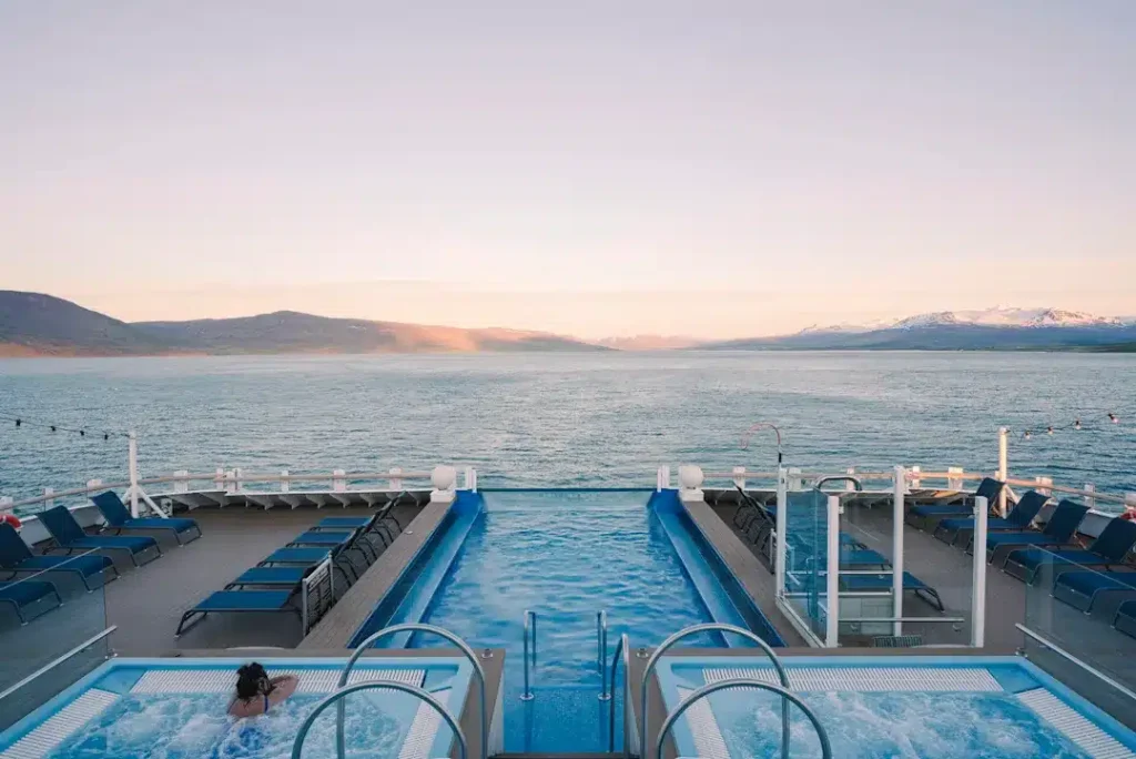 Two ship hot tubs and an infinity pool on an open deck at dusk, with lounge chairs and mountains beyond.