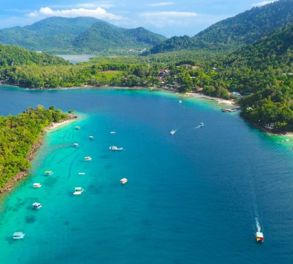 Aerial view of a turquoise bay with boats anchored along a forested peninsula and distant green hills.