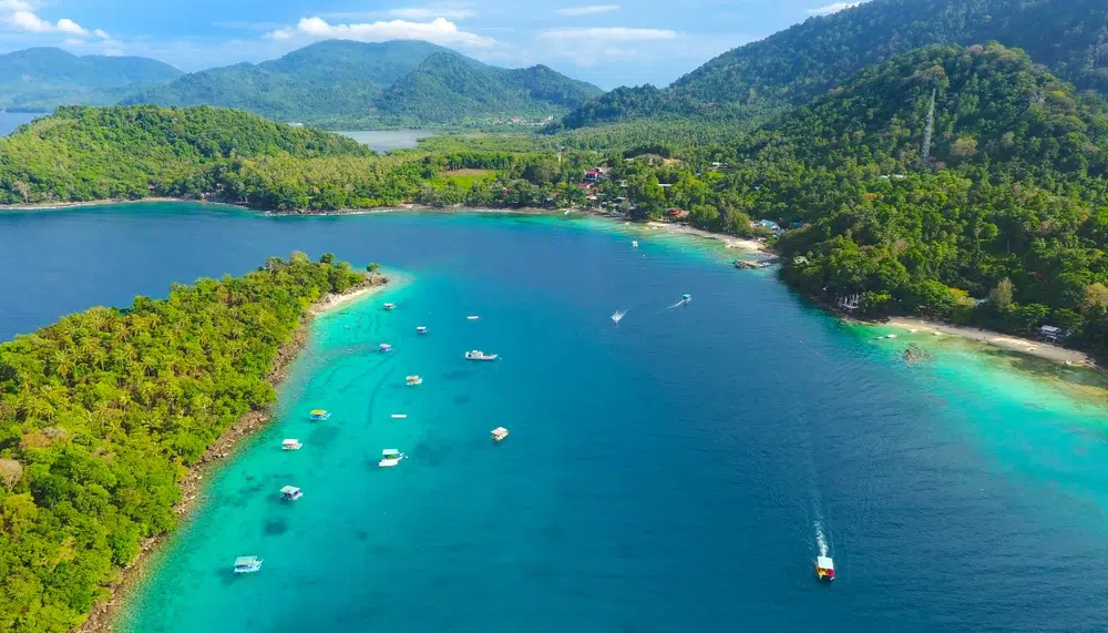 Aerial view of a turquoise bay with boats anchored along a forested peninsula and distant green hills.