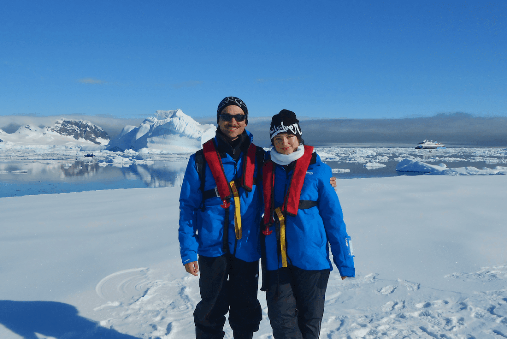 Two smiling people in blue expedition jackets and life vests on a snowy shore with icebergs and a ship behind.
