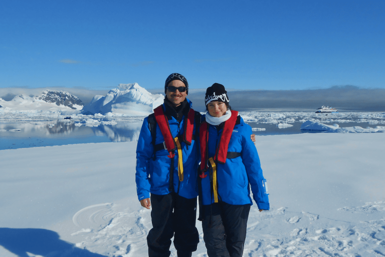 Two smiling people in blue expedition jackets and life vests on a snowy shore with icebergs and a ship behind.