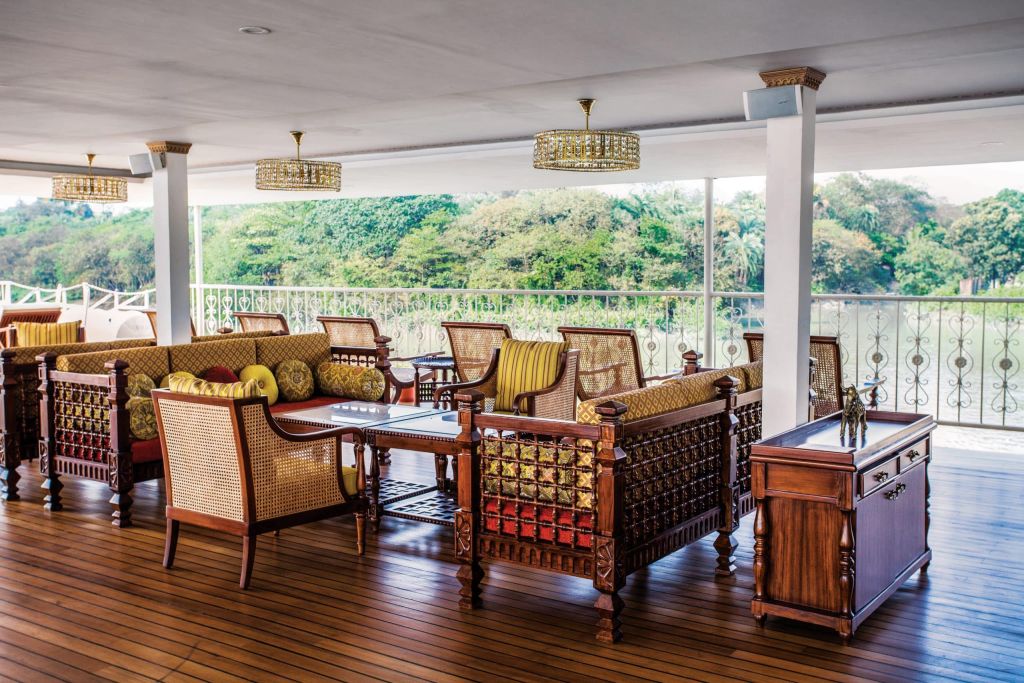 Covered outdoor lounge with carved wooden sofas, yellow cushions, and three ornate gold chandeliers, overlooking a green garden.