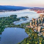 Aerial view of city towers beside a harbor and a forested park with a small lake.