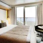 Hotel bedroom with a large bed, neutral bedding, and a balcony door opening to an ocean view.